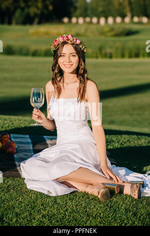 Happy bride en robe blanche et couronne de fleurs assis sur couverture avec verre de vin Banque D'Images