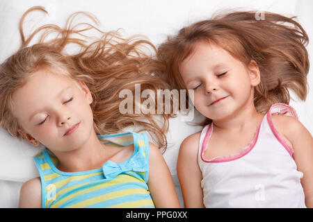 Vue du dessus de l'heureux joli petit filles ou soeurs sourire ensemble, dormir dans le lit, se trouvent étroitement les uns aux autres, voir rêves agréables, vêtus de nightwe Banque D'Images