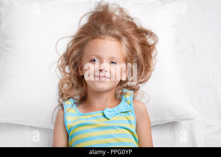 Joli petit gamin avec aspect agréable, les yeux bleus et une peau en bonne santé se penche sur l'appareil photo, pose dans le lit, chambre à coucher a présentation rafraîchie après avoir lo Banque D'Images