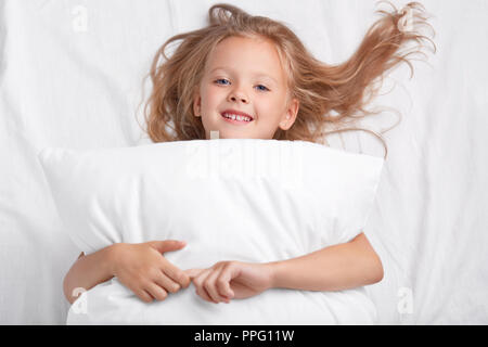 Satisfait playful girl avec un charmant sourire, embrasse un oreiller, se trouve sur l'oreiller blanc, a un bon repos, bénéficie d'éveil, pose dans le lit. Les enfants, reste, rela Banque D'Images