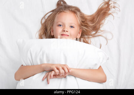 Dreamy blue eyed kid femme aux cheveux clairs, embrasse un oreiller, ressemble à l'appareil photo, pose de façon positive sur le lit blanc, d'être dans la bonne humeur après un sommeil sain. Banque D'Images