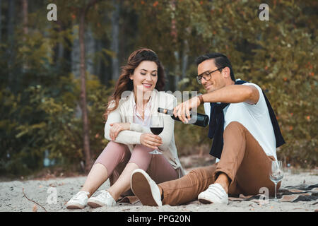 L'homme de verser le vin rouge dans un verre alors qu'elle repose avec femme on sandy beach Banque D'Images