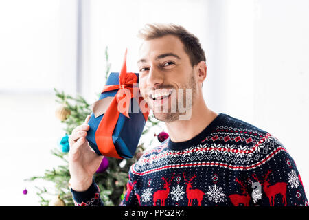 Smiling man in christmas sweater avec boîte-cadeau près de head Banque D'Images