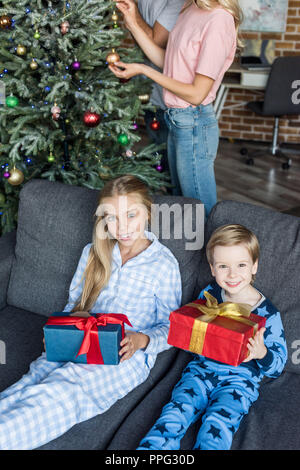 Portrait d'enfants heureux dans pajamas holding christmas gifts and smiling at camera while parents decorating Christmas Tree derrière Banque D'Images