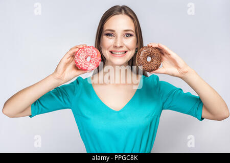 Portrait of smiling young positives belle fille en blouse bleue, tenant debout et montrant le rose et le chocolat donuts avec dents sourire sur bac gris Banque D'Images