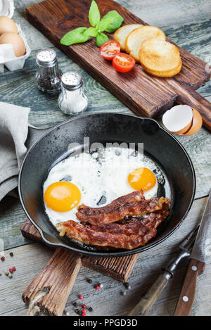 Sur le plat, tranches de bacon, tomate et basilic de moitié dans une poêle en fonte sur une vieille table en bois dans un style rustique Banque D'Images