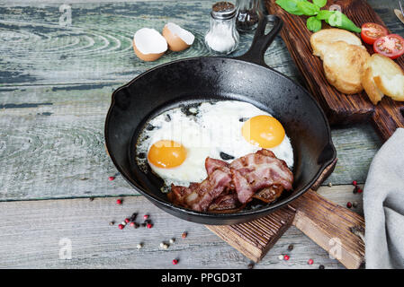 Sur le plat, tranches de bacon, tomate et basilic de moitié dans une poêle en fonte sur une vieille table en bois dans un style rustique Banque D'Images