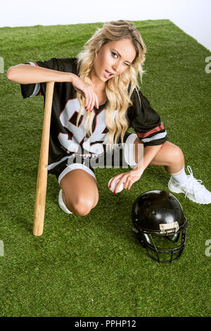 High angle view of athletic jeune femme en uniforme de football américain avec casque et batte de baseball sitting on grass Banque D'Images