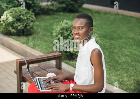 Cheerful attractive élégante african american businesswoman using laptop on bench on street Banque D'Images