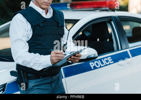 Vue partielle de l'agent de police de sexe masculin d'âge moyen en gilet pare-balles écrit dans le presse-papiers près de voiture à street Banque D'Images