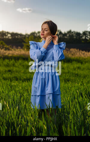 Jeune femme aux yeux clos en bleu et blanc robe rayée est contrôler son earring tout en se tenant dans le domaine de blé vert à un après-midi de printemps Banque D'Images