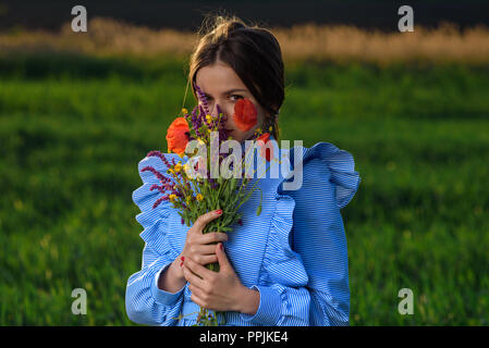 Jeune femme en bleu et blanc robe rayée tient un bouquet de fleurs d'été et à l'intermédiaire de ce dernier, à l'appareil photo tout en se tenant dans le domaine de Banque D'Images