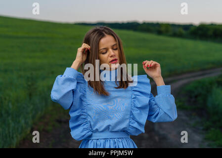 Jeune femme en bleu et blanc robe rayée est debout sur la route de campagne avec ses mains posées près de domaine de blé vert au printemps en soirée. Wom Banque D'Images