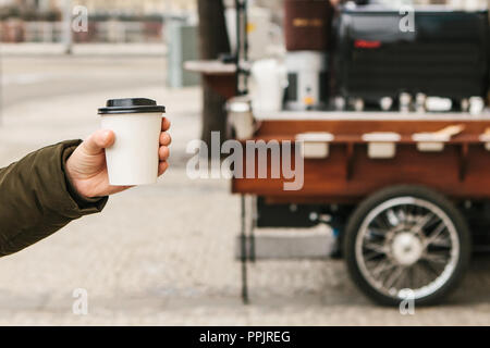 La photographie conceptuelle. La personne est titulaire d'un verre jetable à la main avec du café ou une autre boisson chaude. Street vente de boissons chaudes sur l'arrière-plan. Banque D'Images