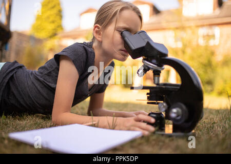Une fillette de 9 ans se trouve sur l'herbe dans le jardin et regarde dans l'oculaire d'un microscope. En face d'elle est une feuille de papier. Banque D'Images