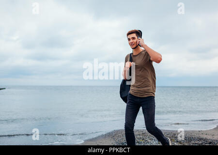 Jeune homme souriant avec guitare acoustique marche sur nuageux beach talking on phone Banque D'Images