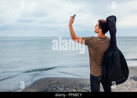 Jeune homme à la guitare acoustique en cas de prendre l'aide de selfies téléphone sur nuageux beach Banque D'Images