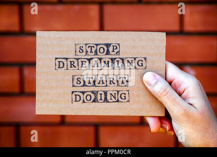 Femme main tenant la carte en carton avec des mots Stop Dreaming Début faisant fait par contre timbres alphabet noir mur arrière-plan. Banque D'Images