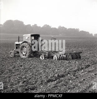 Années 1950, historiques, au milieu d'un terrain accidenté, un agriculteur se dresse sur l'arrière de son tracteur, attaché avec un râteau de sol machine, England, UK. Banque D'Images