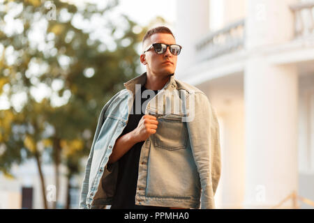 Modèle élégant beau jeune homme avec des lunettes de soleil et veste en jean à la mode dans la ville Banque D'Images