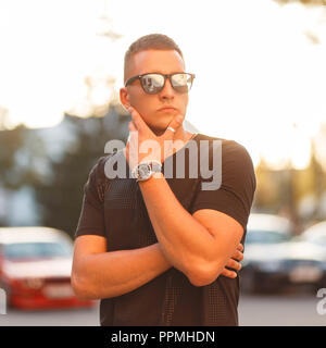Beau jeune homme modèle à la mode avec des lunettes de soleil et en coiffure black fashion T-shirt sur la plage au coucher du soleil Banque D'Images