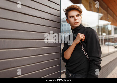 Beau jeune homme dans le quartier branché de vêtements noirs avec un sac pose dans la rue près de mur en bois Banque D'Images