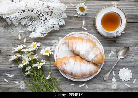 Belle composition : deux croissants, bouquet de fleurs Daisy, tasse de thé et blanc lacy nappes sont sur un vieux fond de bois Banque D'Images