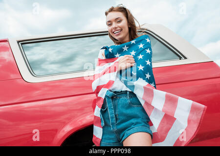 Cheerful girl wrapped in american flag debout près de voiture rouge Banque D'Images