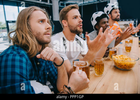 Groupe multiculturel de bouleverser les fans de football masculin en ballon de soccer chapeaux de boire une bière et regarder des match de football à bar Banque D'Images