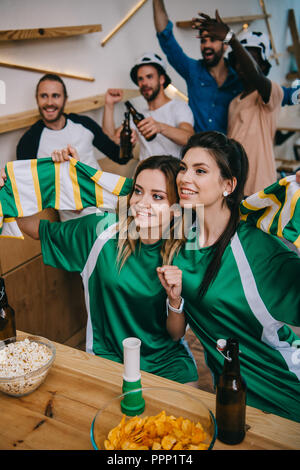 Portrait de la femme en vert fan t-shirts holding foulard et leurs amis masculins debout derrière pendant veille de match de foot au bar Banque D'Images