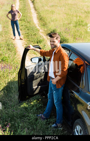 Smiling young man la fermeture de porte de voiture et sa petite amie en attente derrière on rural meadow Banque D'Images