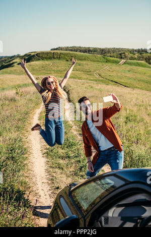 Smiling young woman jumping avec de larges bras pendant que son petit ami en tenant sur smartphone selfies près de voiture sur meadow rural Banque D'Images