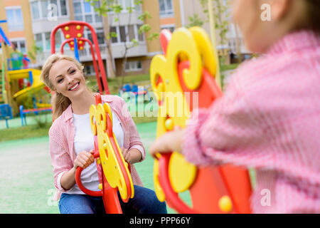 Portrait of Girl riding sur cheval à bascule avec smiling mother à l'aire de jeux Banque D'Images