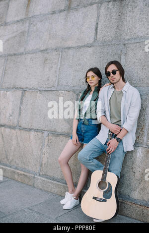 Jeune et heureux couple à lunettes avec guitare par mur en ville Banque D'Images