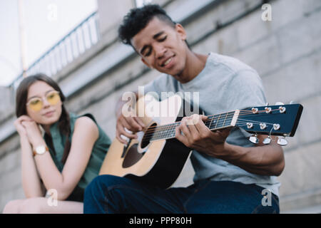 Young woman listening to African American guy à jouer de la guitare à city street Banque D'Images