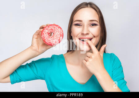 Portrait of young beautiful girl in blue blouse debout, tenant et montrant pink donut, suçant son doigt avec sourire à pleines dents dans la zone de gris Banque D'Images