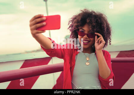 Jolie jeune femme portant des lunettes rouges et en tenant vos autoportraits merveilleux Banque D'Images