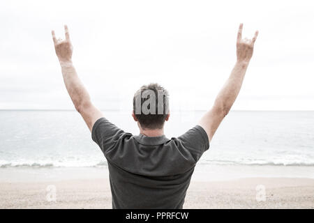 Une vue arrière d'un homme levant les bras en l'air sur une plage déserte et isolée et de faire un signe de cornes radical avec ses mains au mépris Banque D'Images