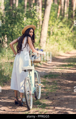 Jeune Femme au chapeau de paille et robe blanche balade à vélo sur le sentier en forêt Banque D'Images