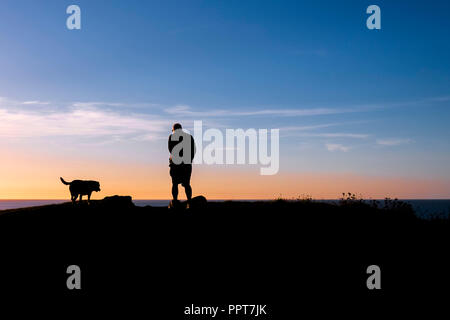 Un homme et son chien vu en silhouette contre un coucher de soleil à Newquay en Cornouailles. Banque D'Images