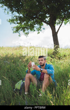 Homme tourist en utilisant smartphone et eating sandwich sur summer meadow with tree Banque D'Images