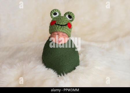 Neuf jours bébé nouveau-né garçon portant un chapeau de grenouille verte. Il est en train de dormir debout alors que dans une emmailloté étirable. Banque D'Images