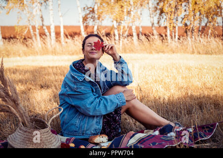 Pique-nique dans l'air frais : une jeune femme en robe et veste en jean tient une feuille rouge et profiter de la nature, assis sur un plaid avec un panier de pique-nique, Banque D'Images