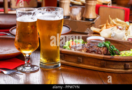 Sex steak avec tomates, salade, courgette, verts et de la sauce sur une planche de bois. Deux verres de bière légère. Tableau de la bière dans le restaurant Banque D'Images