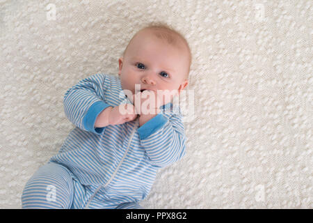 A 2 mois bébé garçon allongé sur le dos sur une couverture blanche. Il porte un pyjama rayé bleu et blanc et a la main dans sa bouche et est à la recherche Banque D'Images