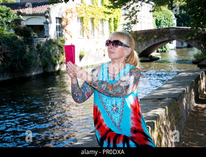 Femme assise seule sur un mur bas à la rive du canal, prenant selfie de son soi et la vue panoramique, ville de Brugge, Belgique. Banque D'Images