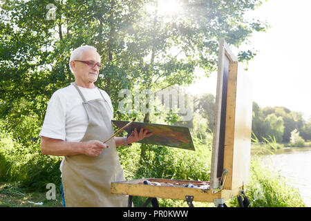 Senior male peintre, comité permanent avec la peinture pinceau et palette de couleurs dans la main, en reprenant la peinture pour photo en rayons de soleil. Bel homme vêtu de blanc T-shirt et un tablier, à la recherche au travail au parc. Banque D'Images