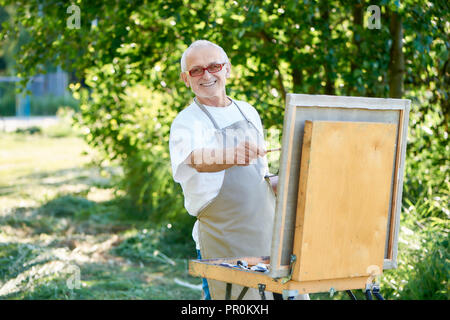 Artiste masculin senior photo dessin peinture à l'aide de parc, palette et chevalet toile. Homme heureux, en portant un tablier, souriant et à la recherche de chevalet, avec photo à l'appareil photo avec finition et chef-d'œuvre. Banque D'Images