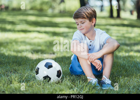 Adorable petit garçon assis sur l'herbe et regardant soccer ball Banque D'Images