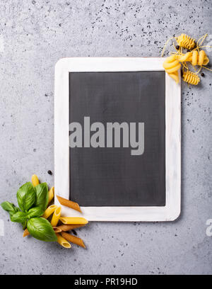 Tableau noir avec grains entiers pâtes italiennes penne, trottole et les feuilles de basilic sur table béton gris. Contexte alimentaire concept pour menu, cafe, resta Banque D'Images